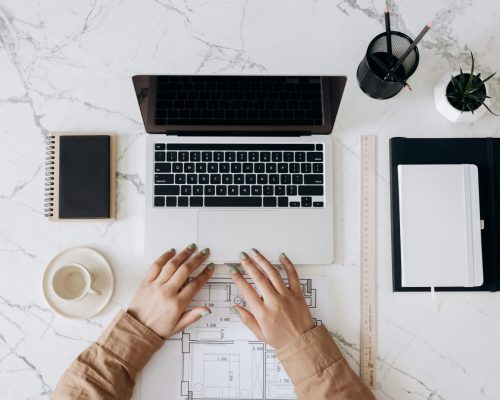 Top view of a stylish home office desk with a laptop, planner, and coffee cup, showing hands on a blueprint.